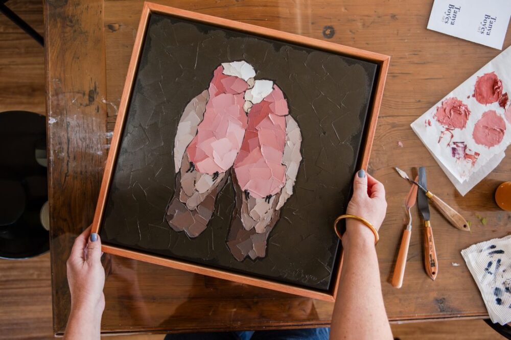 Overhead view of artist Tanya Boyes holding a framed palette knife painting of two pink galahs on a dark background