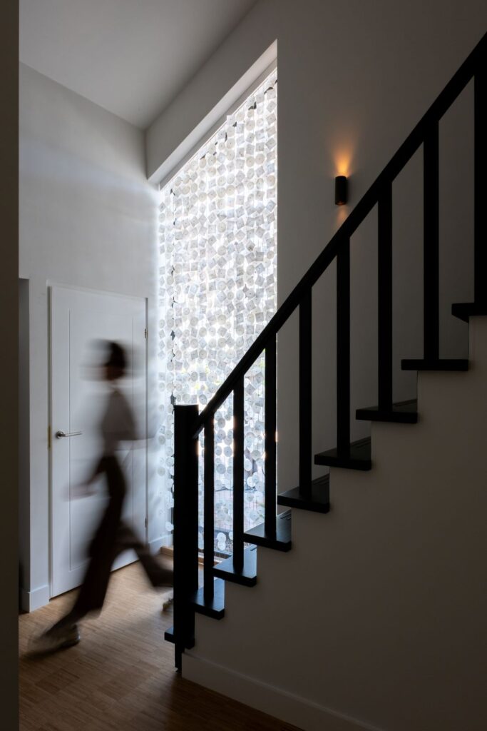Tall translucent installation filtering daylight beside a black staircase as a blurred figure walks past