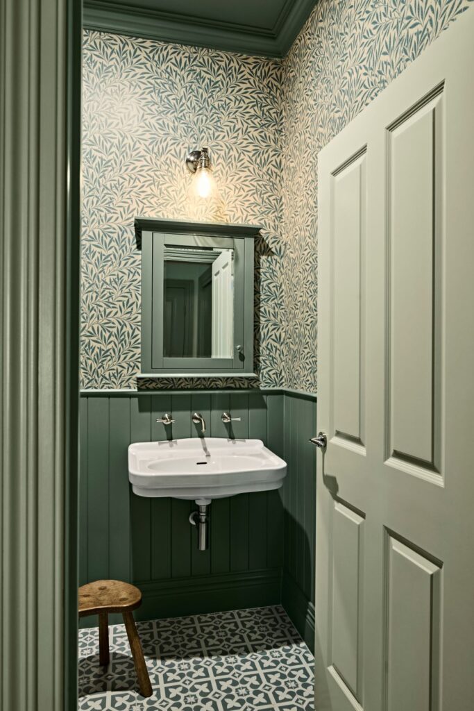 Country-style powder room with blue botanical wallpaper, deep green wall panelling, a white basin and patterned floor tiles
