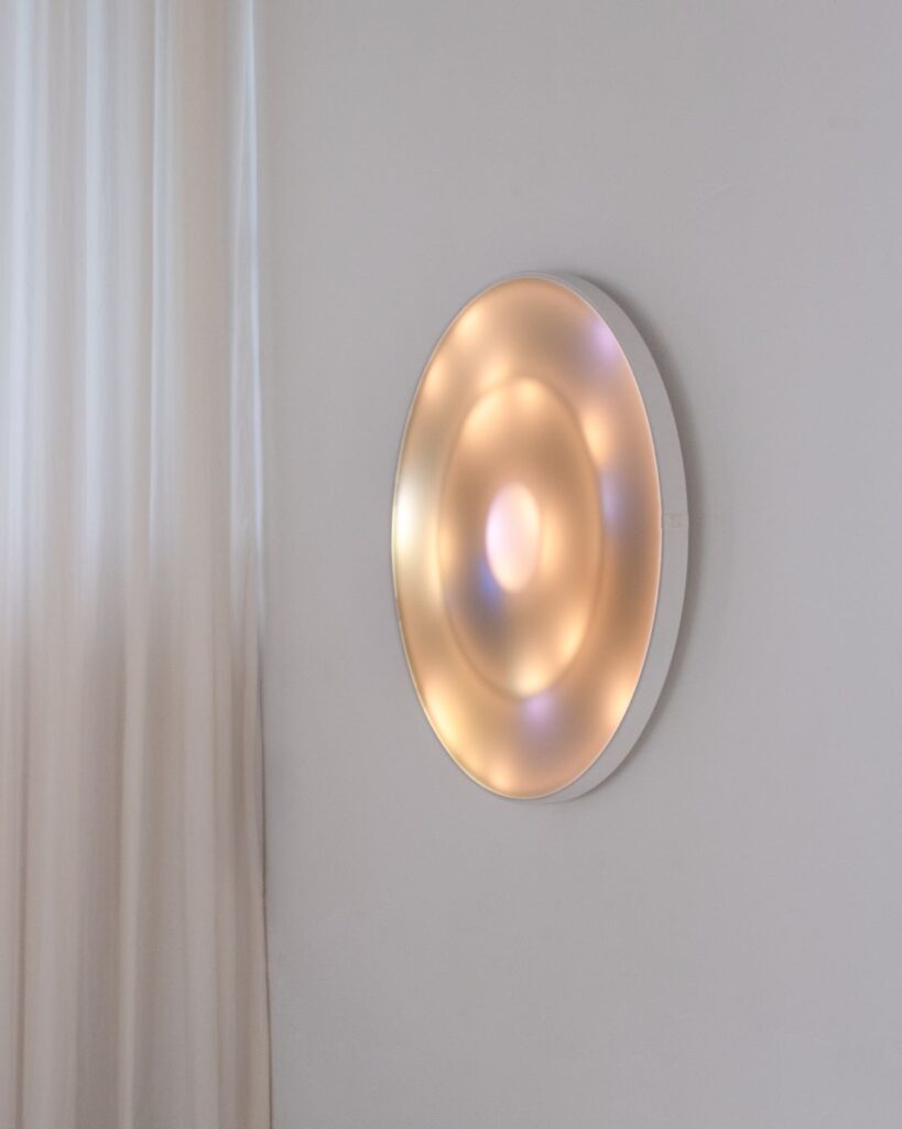 Circular wall-mounted light artwork glowing in warm amber tones beside soft sheer curtains
