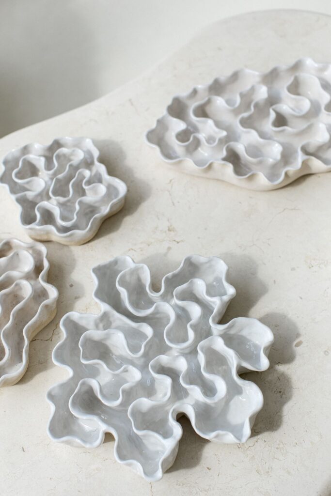 Glossy white ceramic sculptures with layered, undulating forms arranged on a pale stone surface.