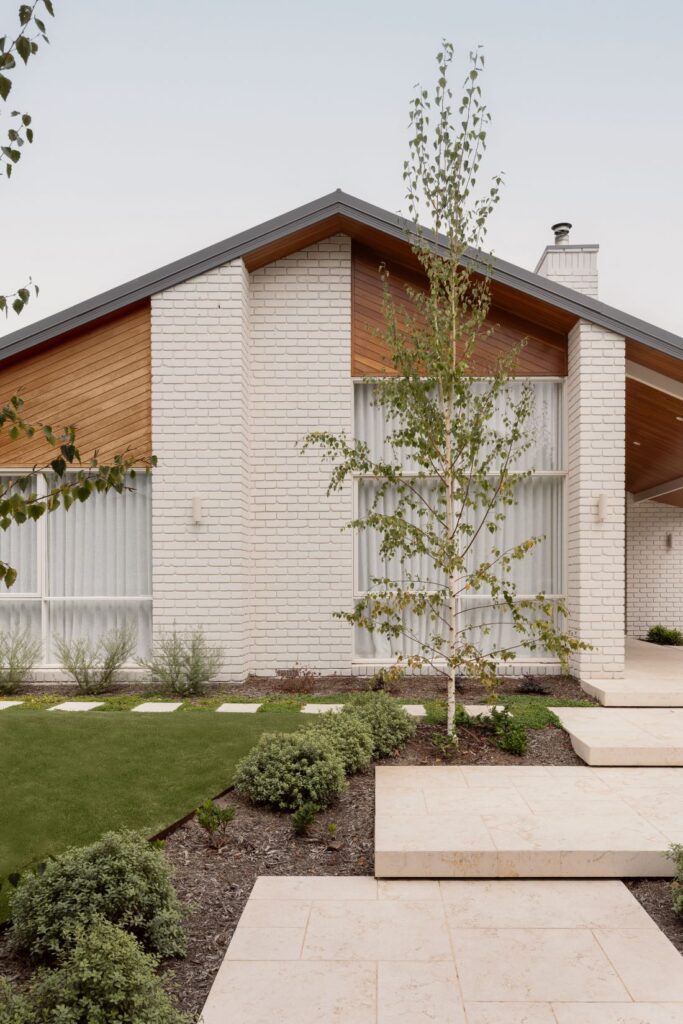 White brick home exterior with warm timber cladding, a gabled roof, large windows, landscaped garden beds and pale stone stepping paths.