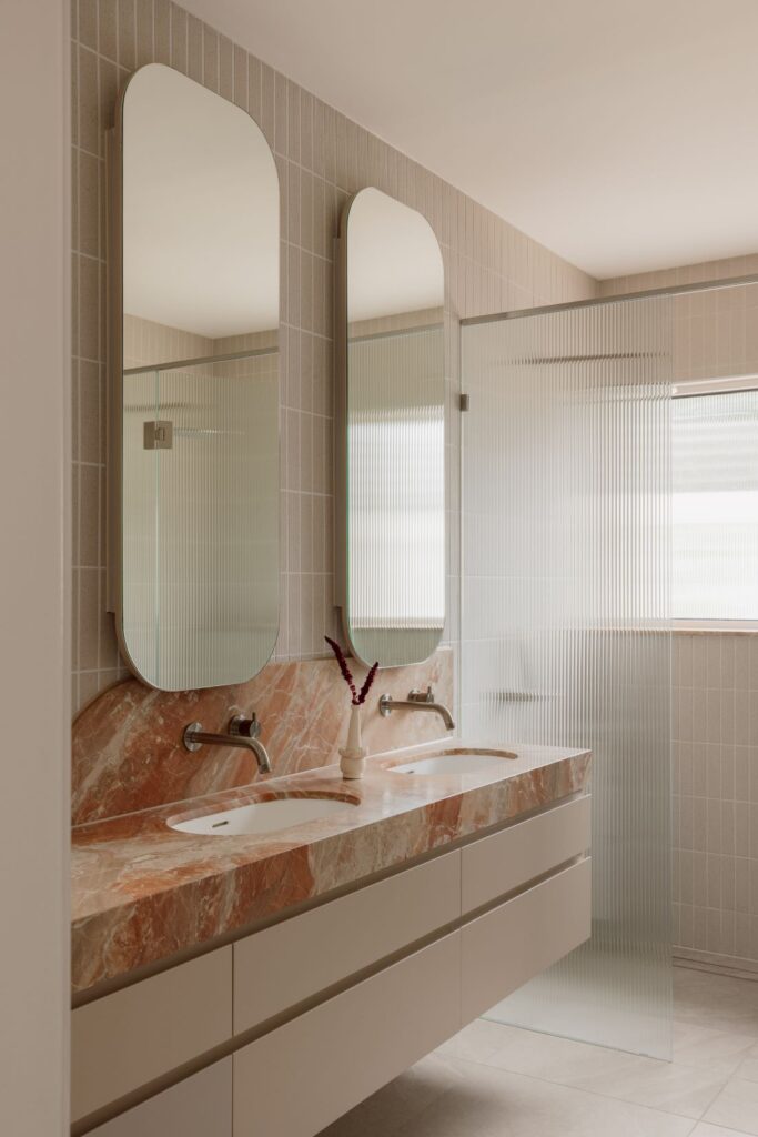 Hampton bathroom with peach-toned marble double vanity, arched mirrors, brushed metal tapware and textured wall tiles.