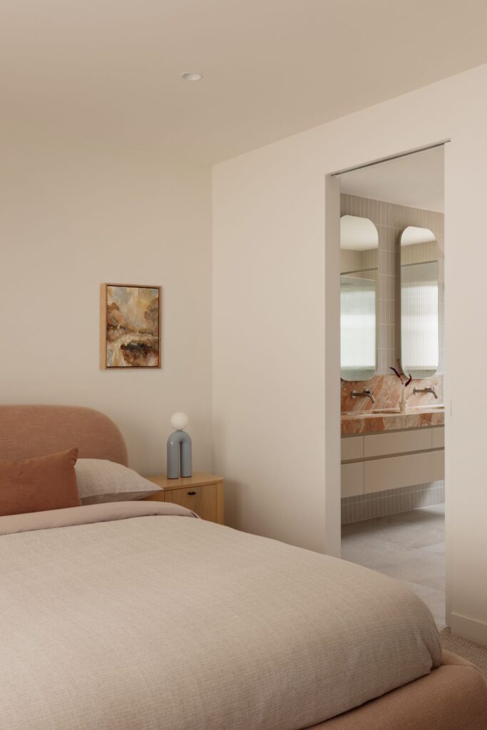 Hampton bedroom with soft neutral bedding, curved upholstered bedhead and view into a peach-toned marble ensuite.
