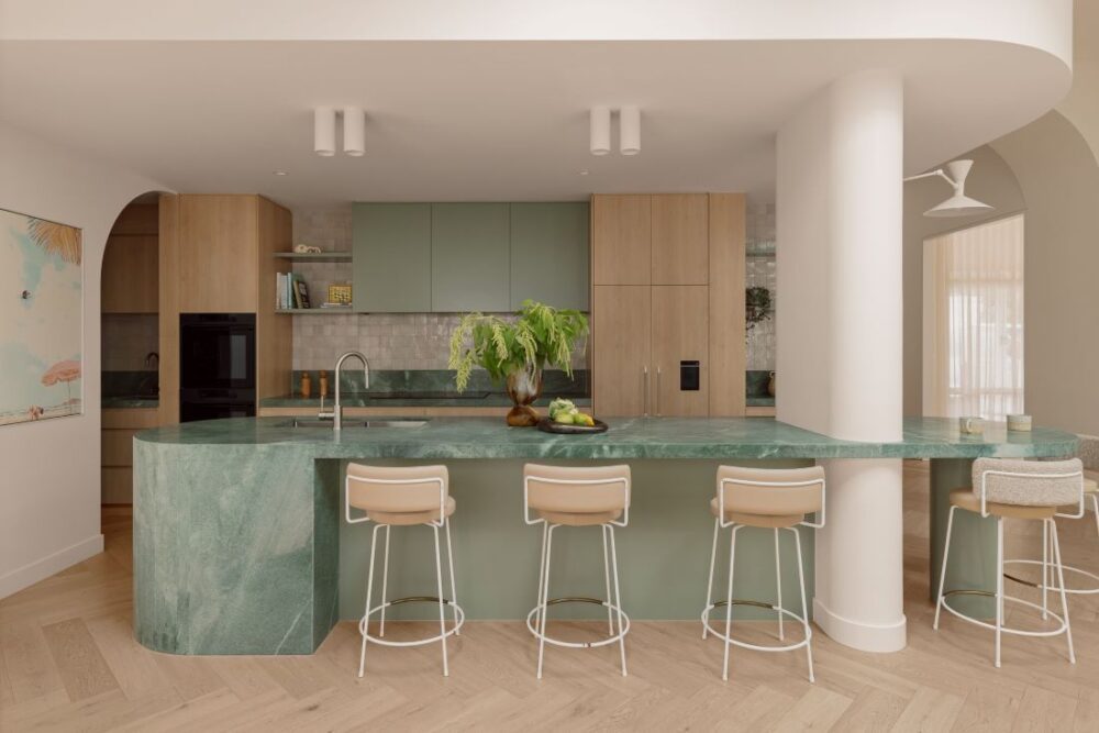 Hampton kitchen with a curved green quartzite island, pale timber cabinetry, arched ceiling details and herringbone timber flooring.