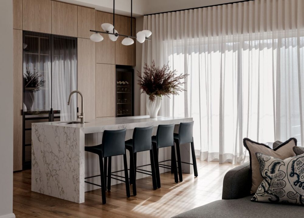 Contemporary kitchen with a marble waterfall island, four dark bar stools, light timber cabinetry, sculptural pendant light and full-height sheer curtains