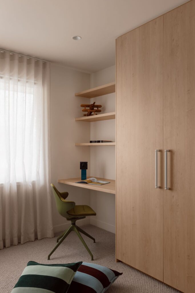Built-in study nook with pale timber desk and shelving, floor-to-ceiling wardrobe, green chair, sheer curtains and neutral carpet.
