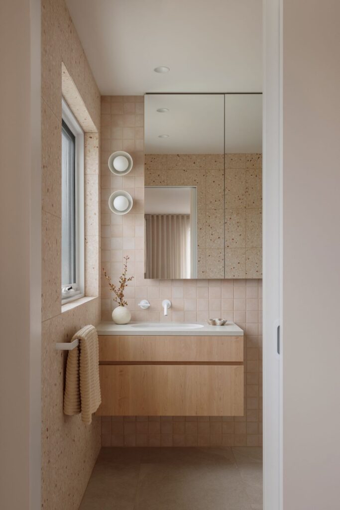 Peach-toned bathroom with square wall tiles, pale timber floating vanity, mirrored cabinetry, round wall lights and a terrazzo-framed window.