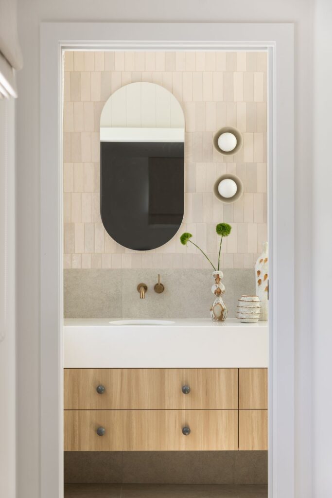 Waterhousia bathroom featuring an arched mirror, cream tiled wall, timber vanity, brass tapware and soft neutral styling details.