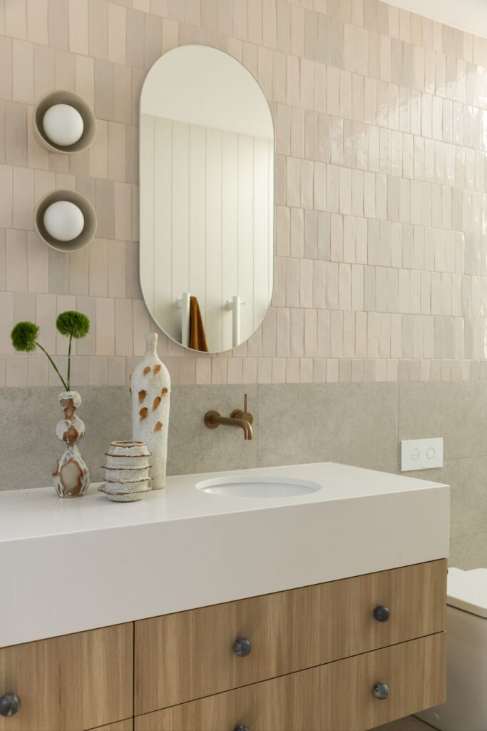 Waterhousia bathroom with cream textured wall tiles, curved mirror, timber vanity, brass tapware and neutral decorative accessories.