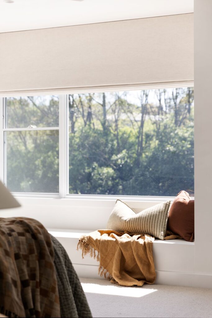 Waterhousia bedroom with a built-in window seat, neutral and rust cushions, ochre throw and large window overlooking leafy greenery.