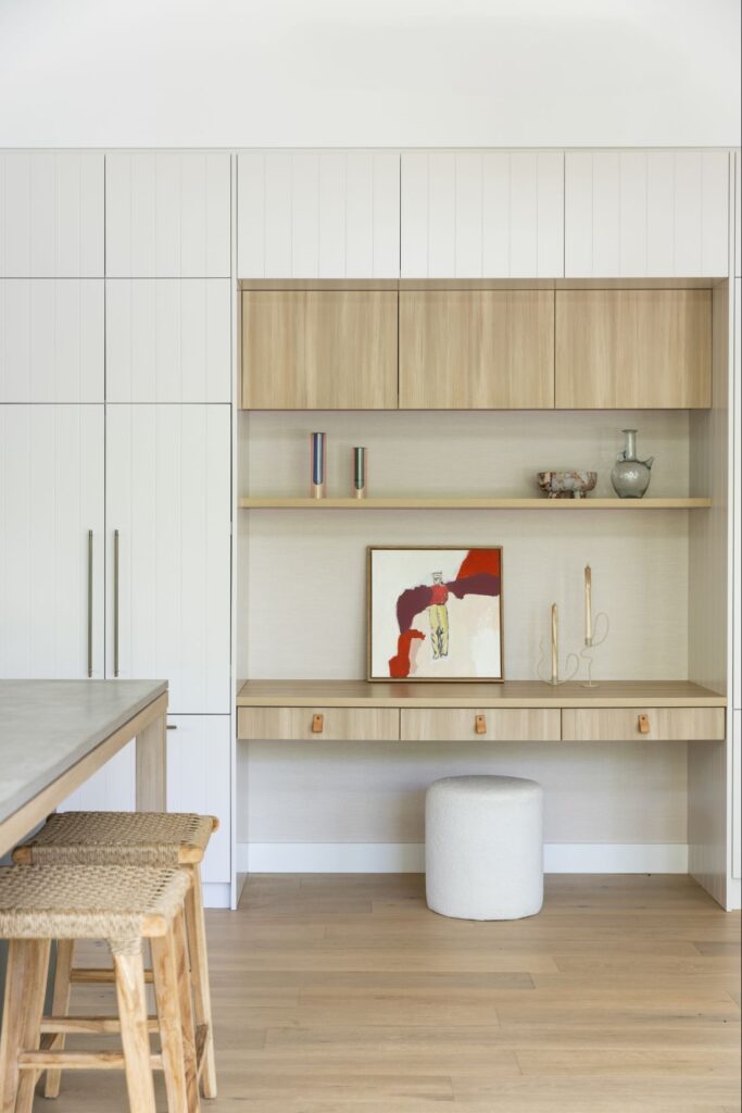 Waterhousia kitchen featuring built-in white cabinetry, timber shelving, integrated desk nook, artwork, woven stools and neutral styling.