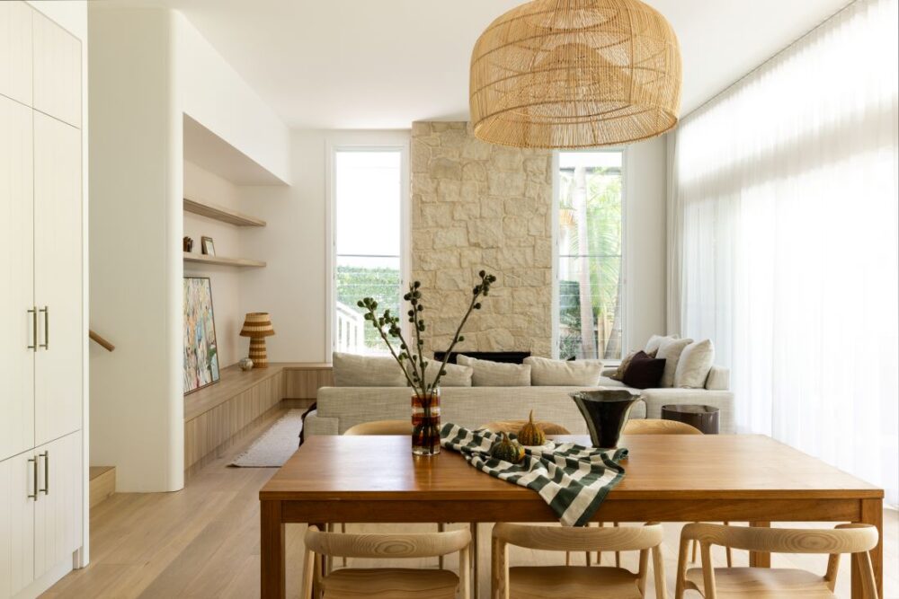 Waterhousia open-plan living and dining area featuring a timber dining table, woven pendant light, neutral sofa, built-in shelving and stone fireplace.