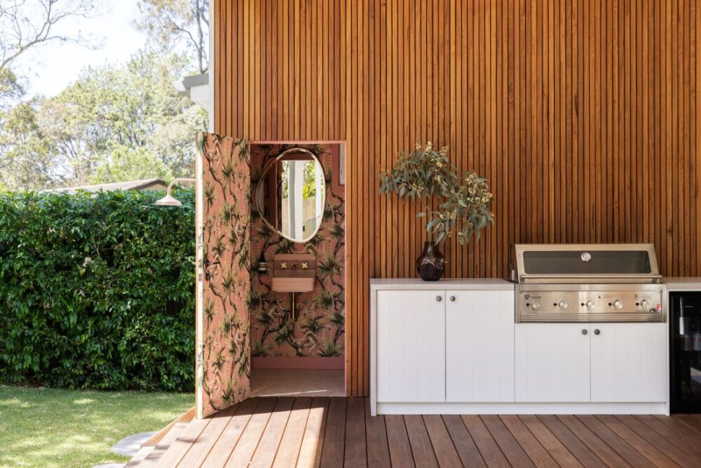 Alt Text: Waterhousia outdoor kitchen featuring timber wall cladding, white cabinetry, built-in barbecue, timber decking and a patterned doorway beside a garden.