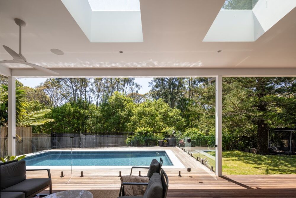 Alt Text: Waterhousia outdoor living area featuring a covered alfresco space, swimming pool, timber decking, glass balustrade and leafy bushland views.