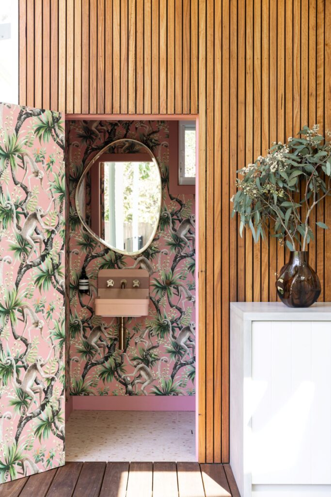 Waterhousia outdoor powder room featuring pink botanical wallpaper, round mirror, blush basin, timber wall cladding and white cabinetry.