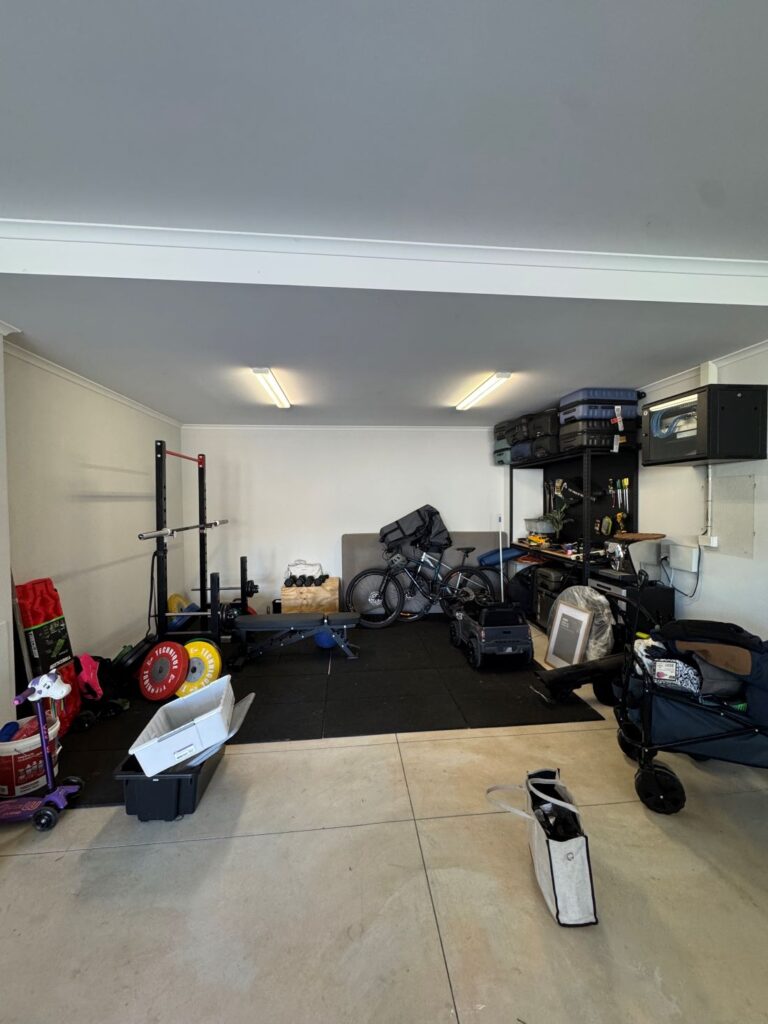 Garage before organisation with exercise equipment, bikes, storage boxes and household items spread across the floor and shelving.