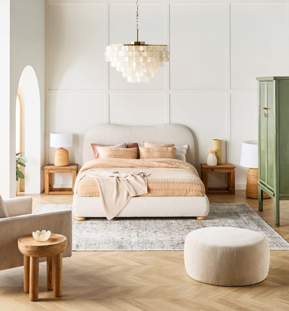 Neutral bedroom featuring an upholstered bed, matching wooden bedside tables, table lamps, pendant light, rug and soft beige décor.