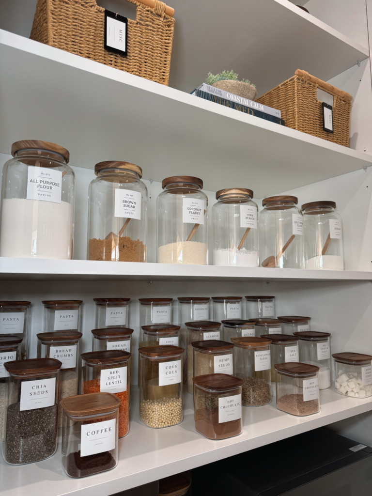 Organised pantry featuring labelled glass jars, food containers, woven baskets and neatly arranged kitchen staples on white shelves.