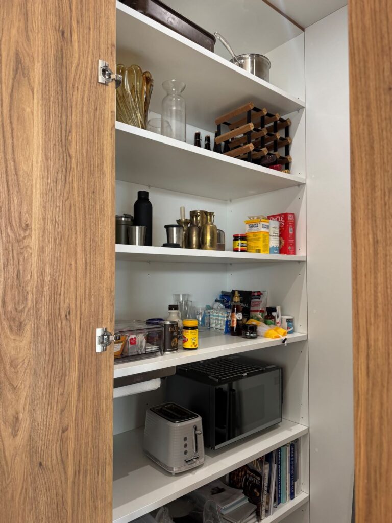 Pantry before organisation with open shelves holding kitchen appliances, bottles, jars, containers and household items.