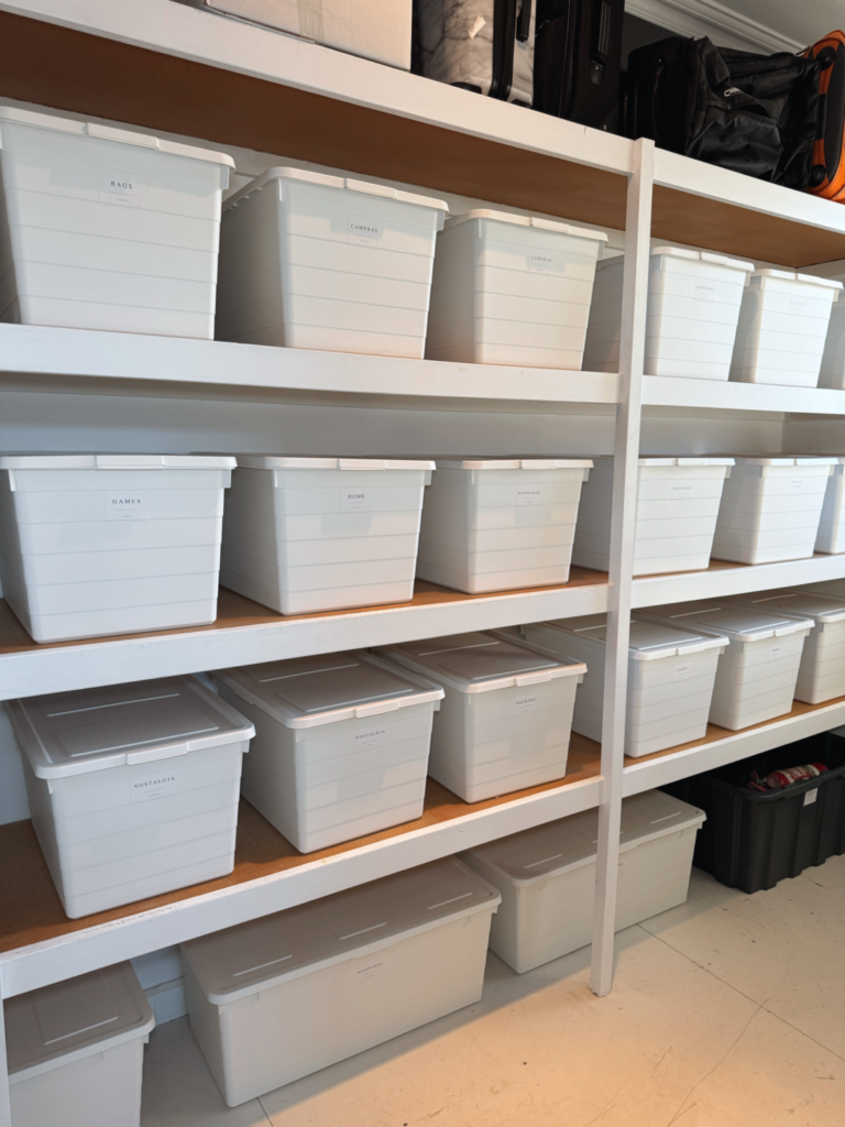 Organised storage room featuring white labelled storage bins neatly arranged on shelving with luggage and containers stored above.