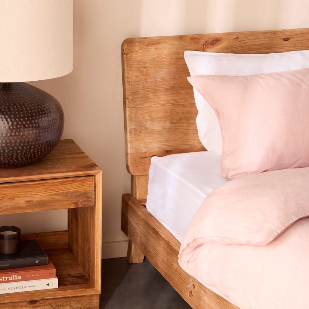 Close-up of a timber bed frame beside a wooden bedside table with a lamp, books and soft pink bedding in a warm neutral bedroom.