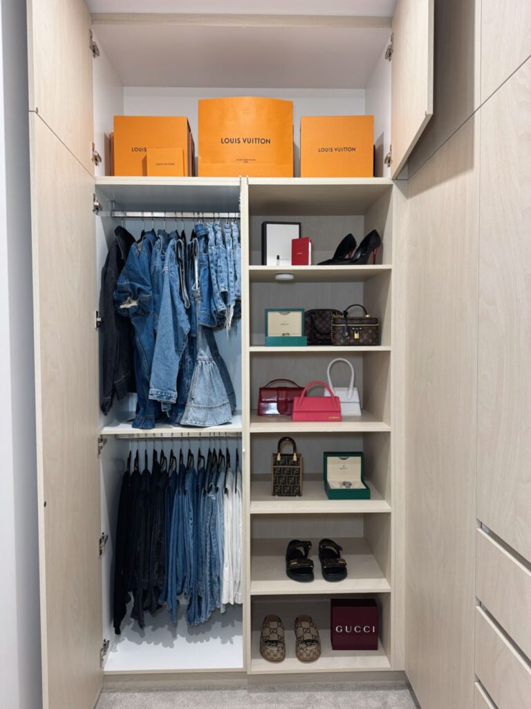 Organised walk-in wardrobe featuring neatly hung clothing, shelves displaying handbags and shoes, labelled luxury storage boxes and built-in cabinetry.