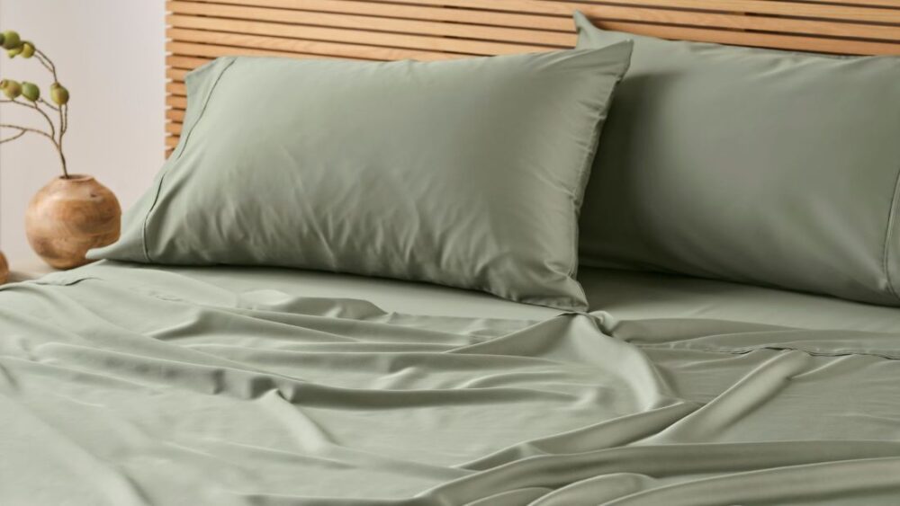 Close-up of a warm minimalist bedroom featuring sage green bedding, soft layered textiles, matching pillows, a timber slatted bedhead and natural bedside styling.
