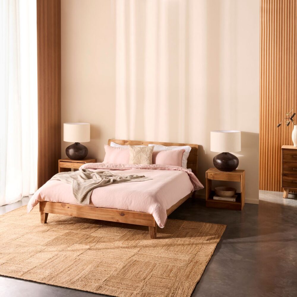 Bedroom with a timber bed, matching bedside tables, table lamps, soft pink bedding and a large woven rug in a warm neutral interior.