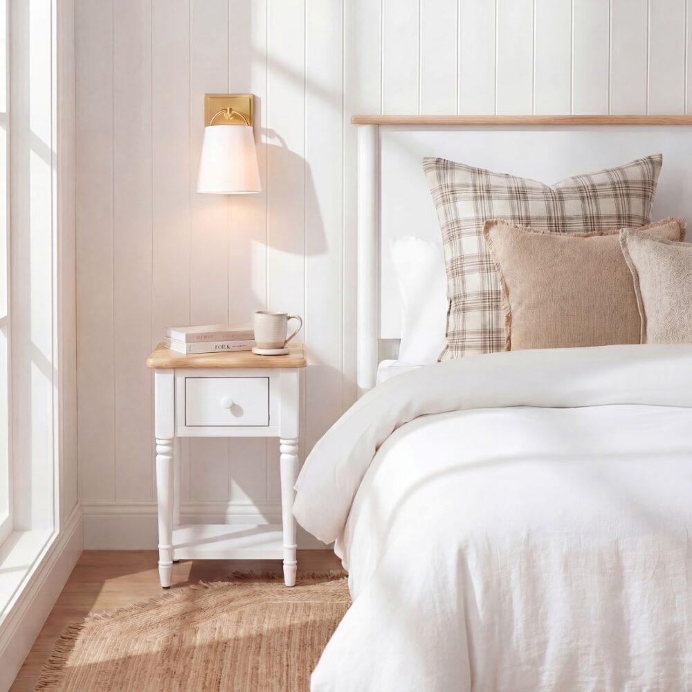 Bright bedroom corner featuring a white bedside table with wooden top, wall lamp, books, cup, white bed, neutral pillows and woven rug.